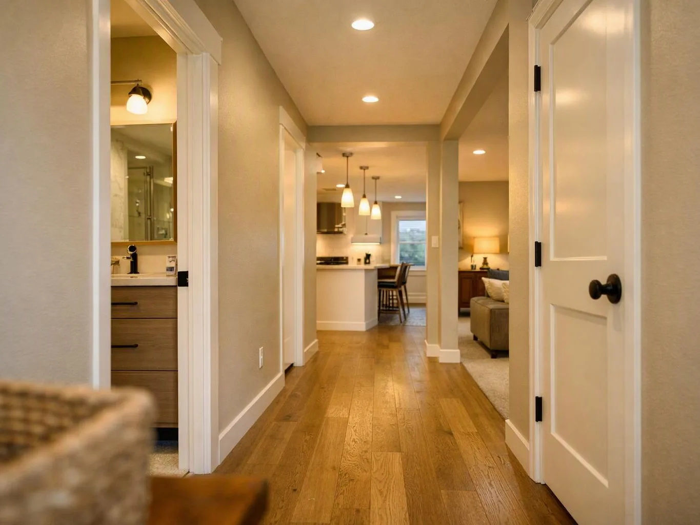 A bright hallway with wood floors leads past open doors to a bathroom, kitchen, and living area