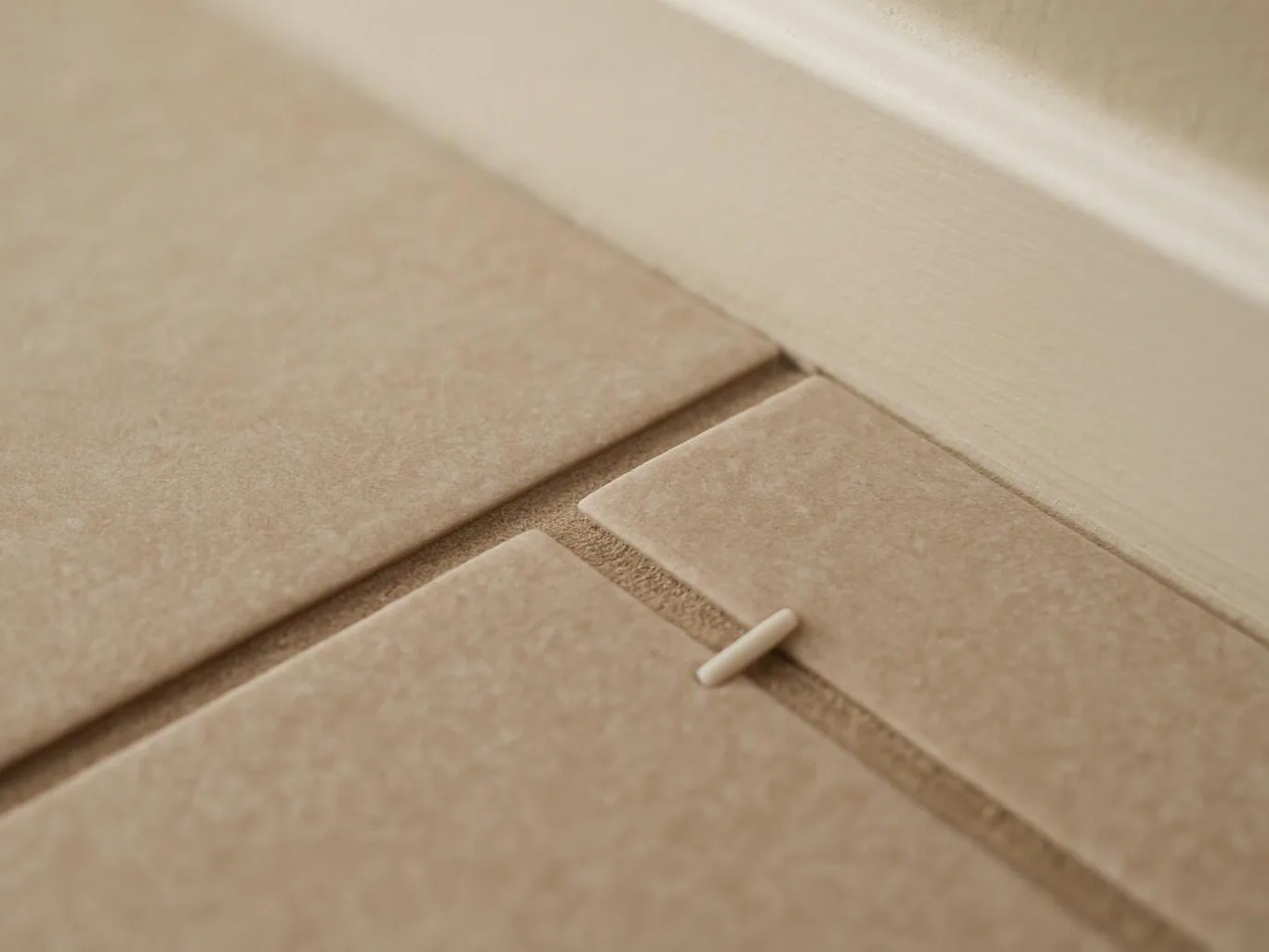 Close-up of beige floor tiles meeting a white baseboard, with a small plastic spacer between the seams