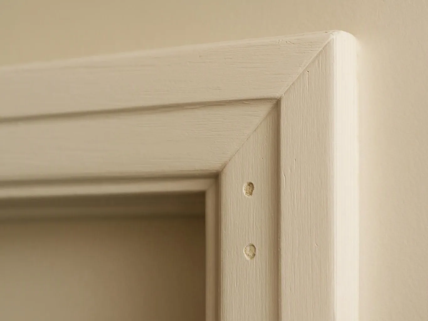 Close-up of a beige wooden door frame corner with visible screw holes on a plain wall