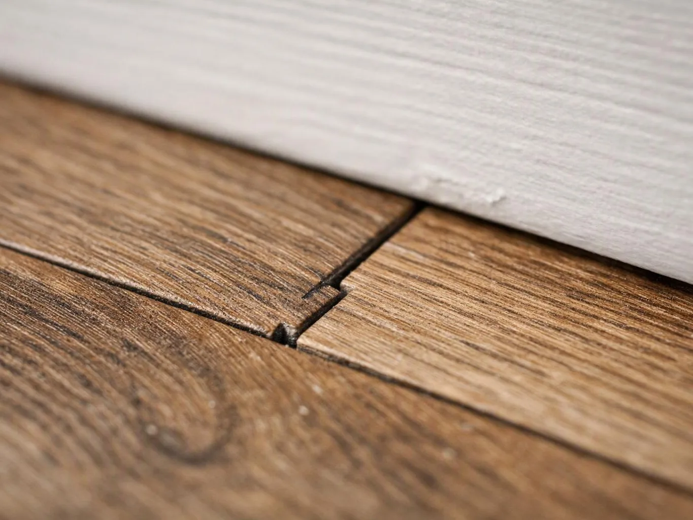 Close-up of a wooden floorboard seam next to a white baseboard, showing a narrow gap and slight damage