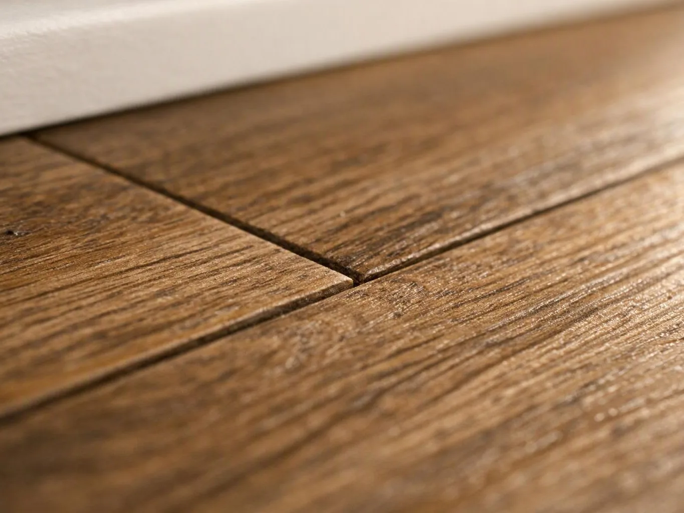 Close-up of a wooden floorboard seam with a small gap and blurred wall above