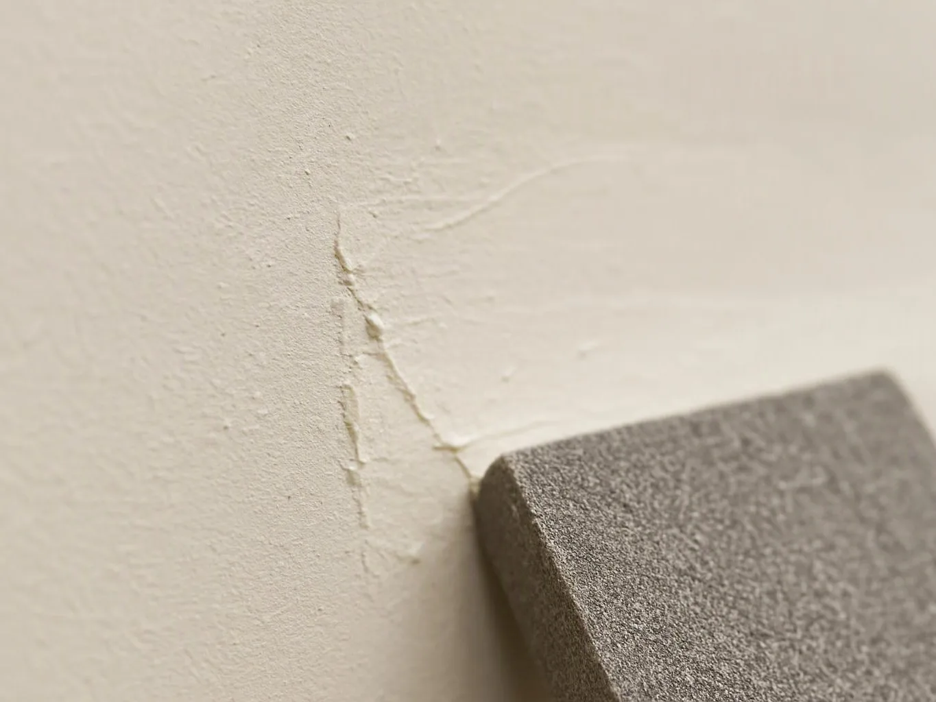 A gray sanding block touches a cracked, peeling painted wall in close-up