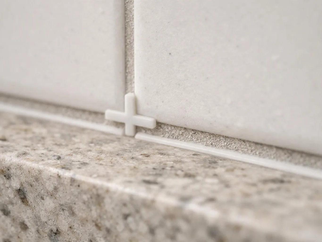 A white tile spacer sits between two tiled walls above a speckled granite ledge
